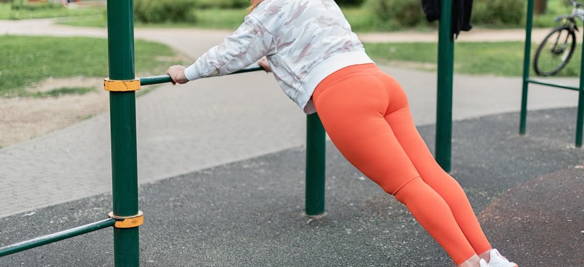 A woman in sportswear performs exercises on outdoor fitness equipment.