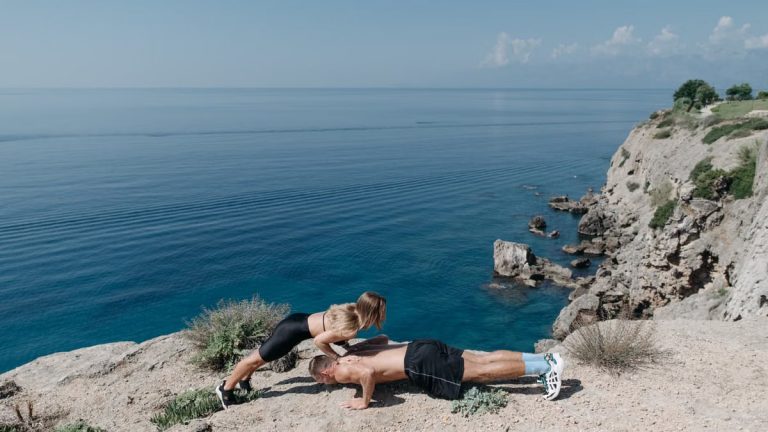 A couple performs a partner workout on a scenic cliffside, overlooking the ocean.